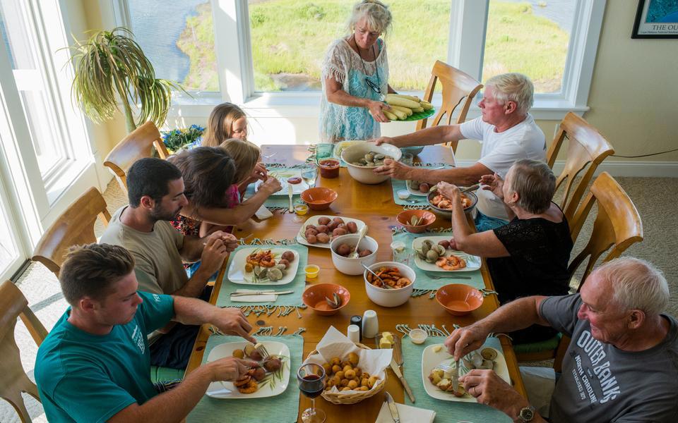 High angle view of a family around a large dining table covered with seafood. Windows reveal the sound just outside.
