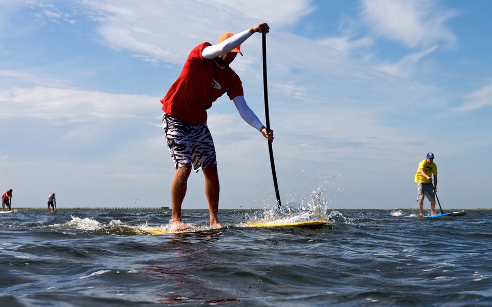 Man in a red shirt paddles hard on his yellow paddleboard while others paddle their SUPs in the background
