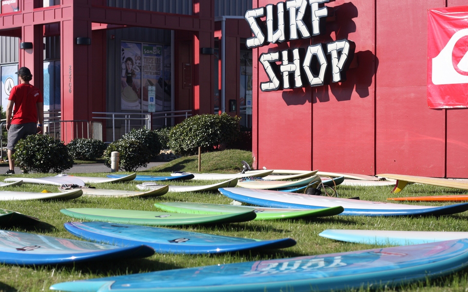 A dozen fiberglass surfboard rest on the grass in front of the red exterior of Real Kiteboarding with the Surf Shop sign