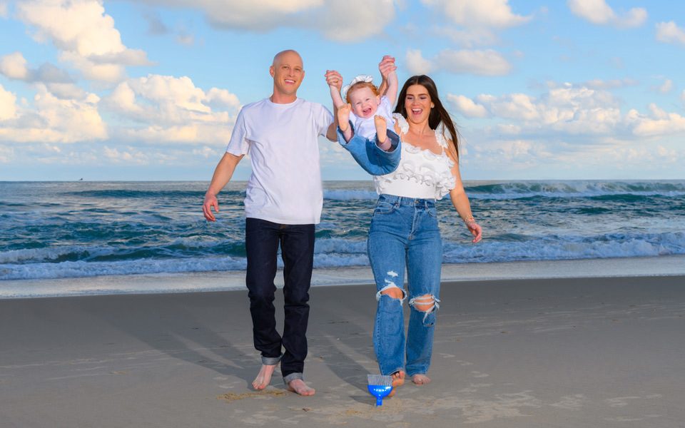 Young couple in white shirts, blue jeans, and bare feet swing a toddler between them walking on the beach by the ocean