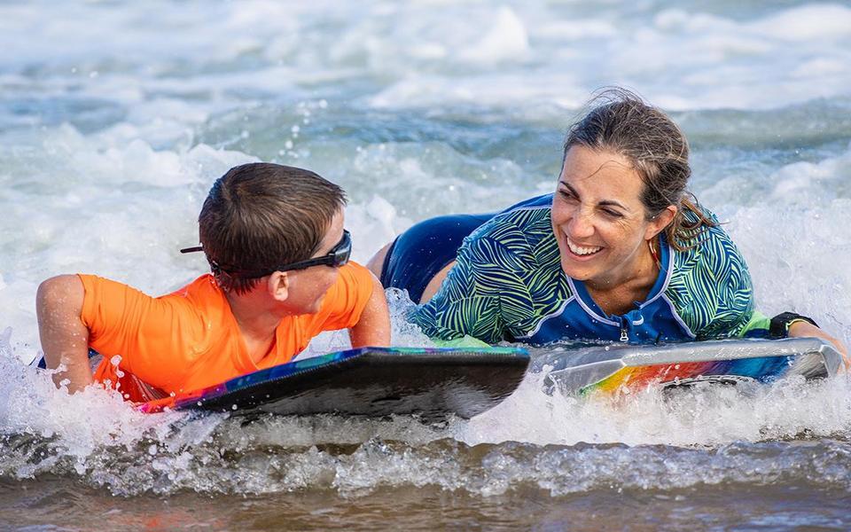 Adult and child smile widely at each other as they ride body boards up the beach in the shallow water