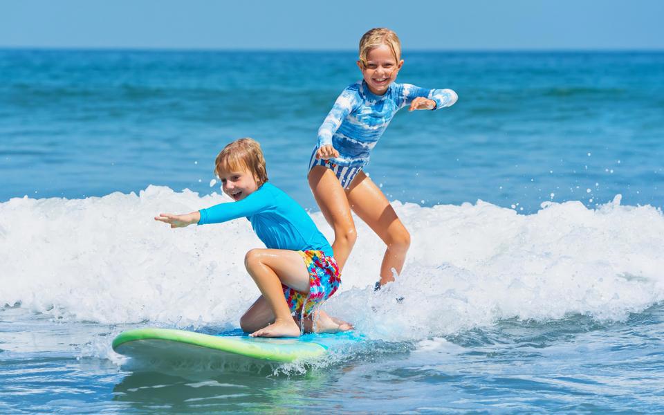 Two young girls ride as small wave on a longboard surfboard with big grins on their faces