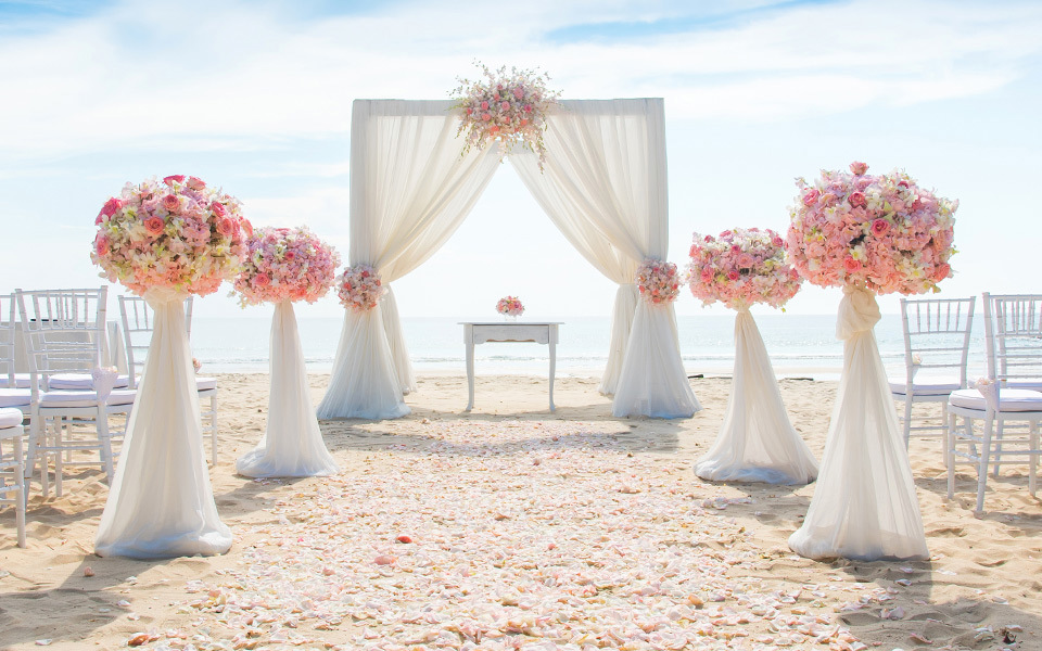 Pink floral arrangements line an aisle looking leading to a gauze-draped wedding alter on the beach.