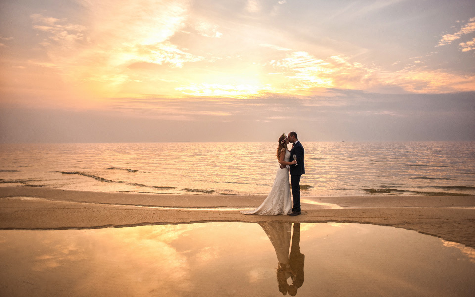 Bride in a wedding dress kisses her man in a suit standing on the sand at sunrise between a reflecting tidal pool and the sea