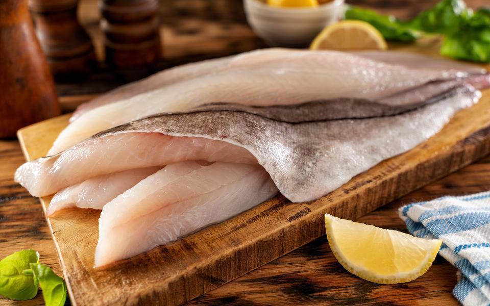 Fresh fillet of white fish rest on a cutting board with a slice of lemon and greenery nearby