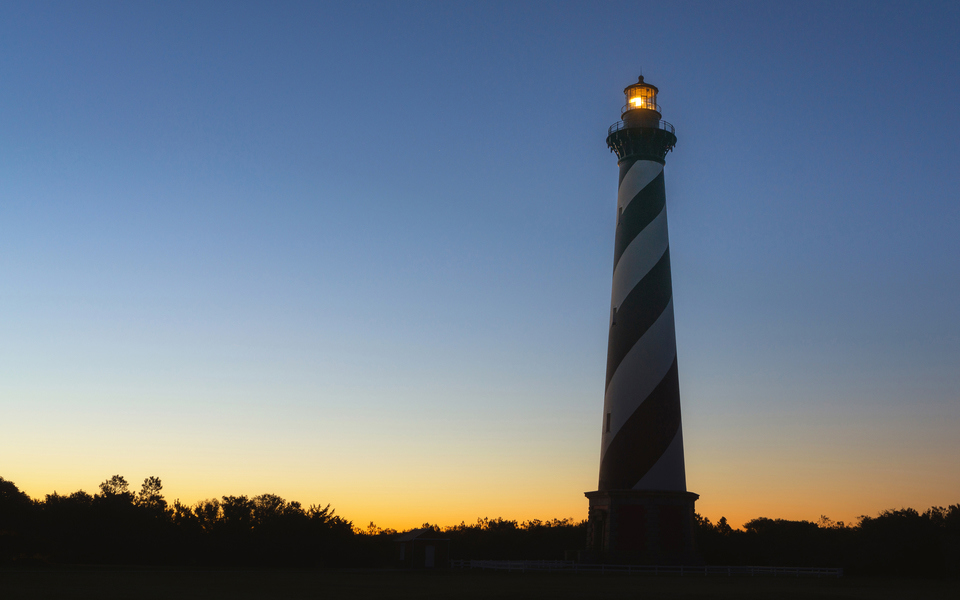 Cape Hatteras Lighthouse mostly silhouetted by a clear twilight sky, dark blue fading down to orange at the treeline