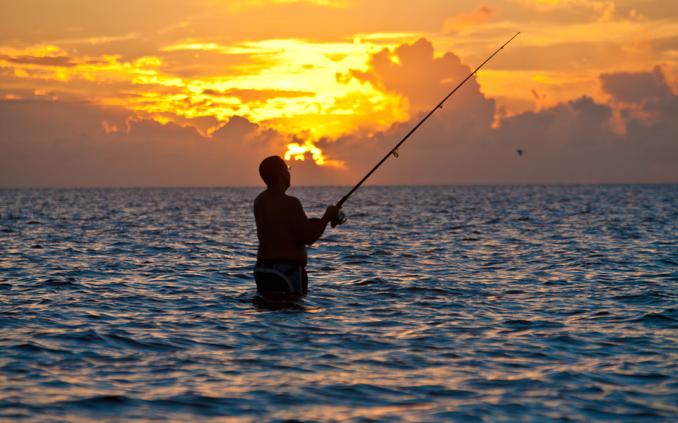 Silhouette of a person surf fishing standing waist deep in the ocean in front of a gorgeous orange sunrise framed in clouds