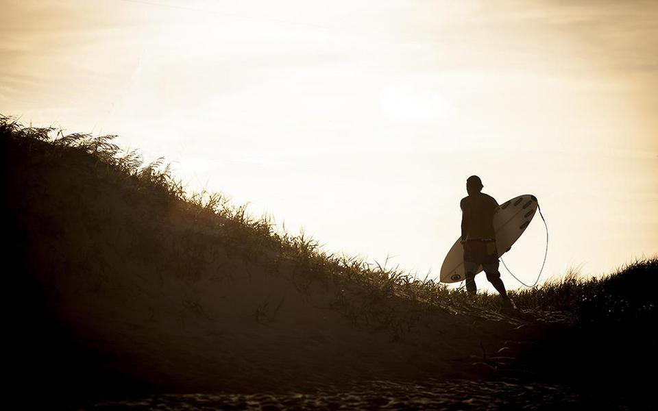 Surfer with surfboard under an arm traverses a grassy due in silhouette heading toward the ocean
