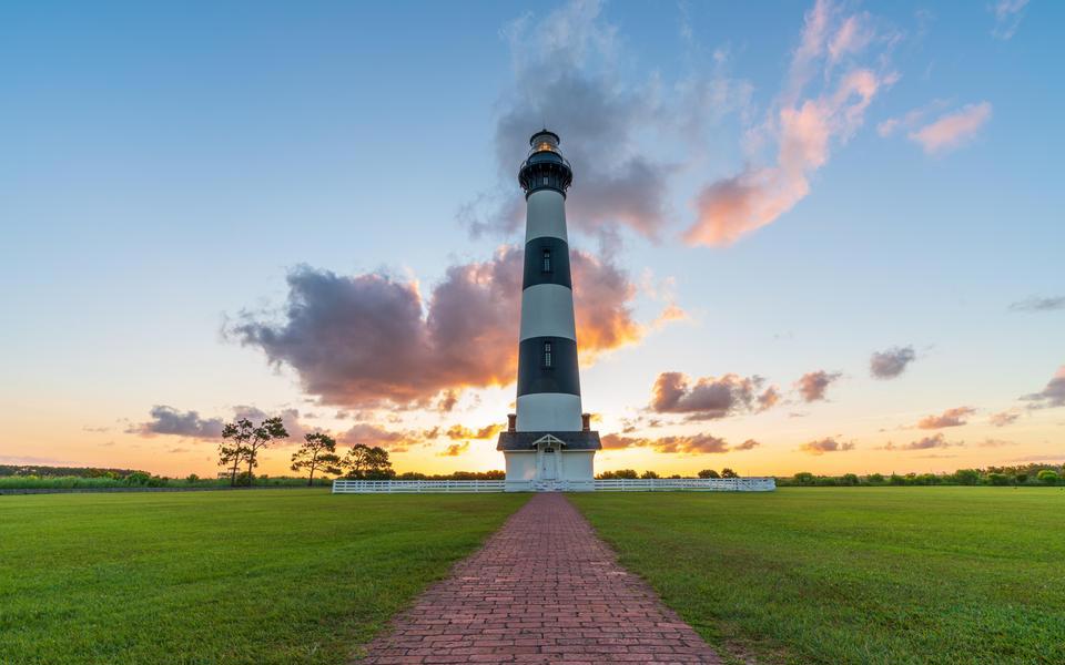 Bodie Island Lighthouse stands tall at the end of a red brick wall flanked by green grass under a blue sky with pink clouds