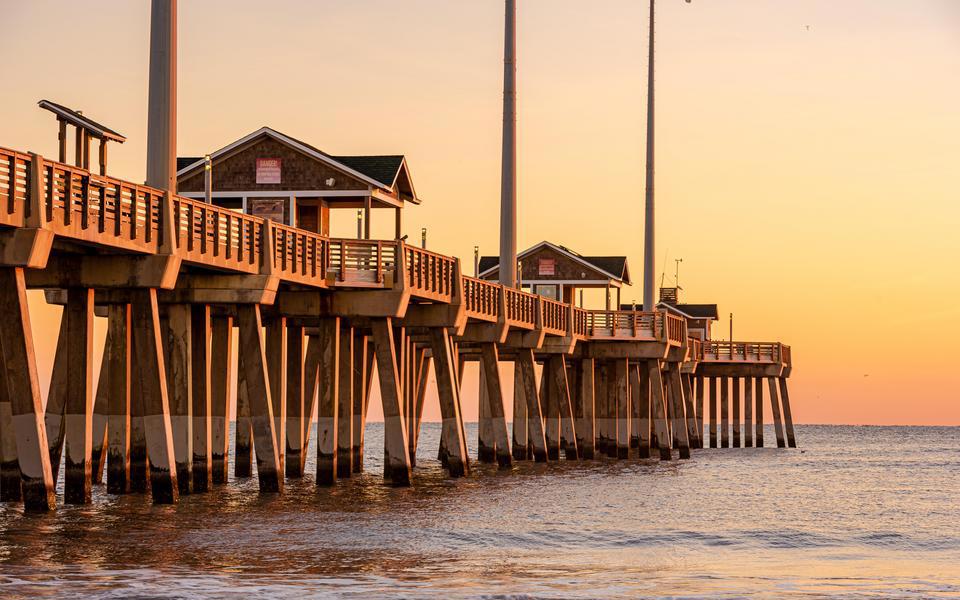 Jennette's Pier, seen from the beach on the south side, is bathed in orange light of sunrise against a pink and orange sky
