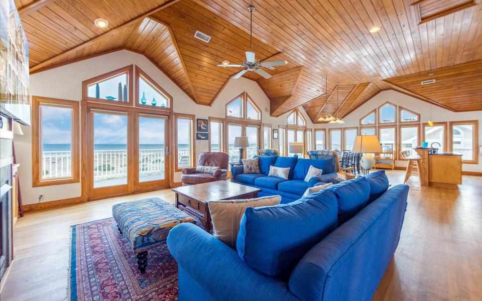 Warn natural wood ceiling spans a large vacation home great room with blue couches and a wall of windows to the sea