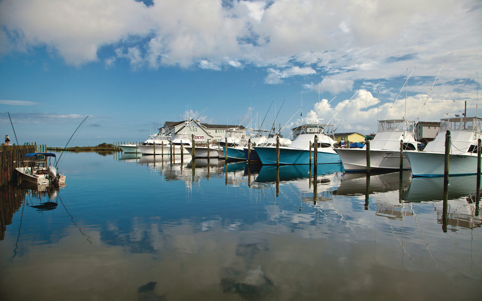Slick calm water reflects the skies, puffy clouds, and colorful off-shore charter fishing boats in Hatteras Island marina