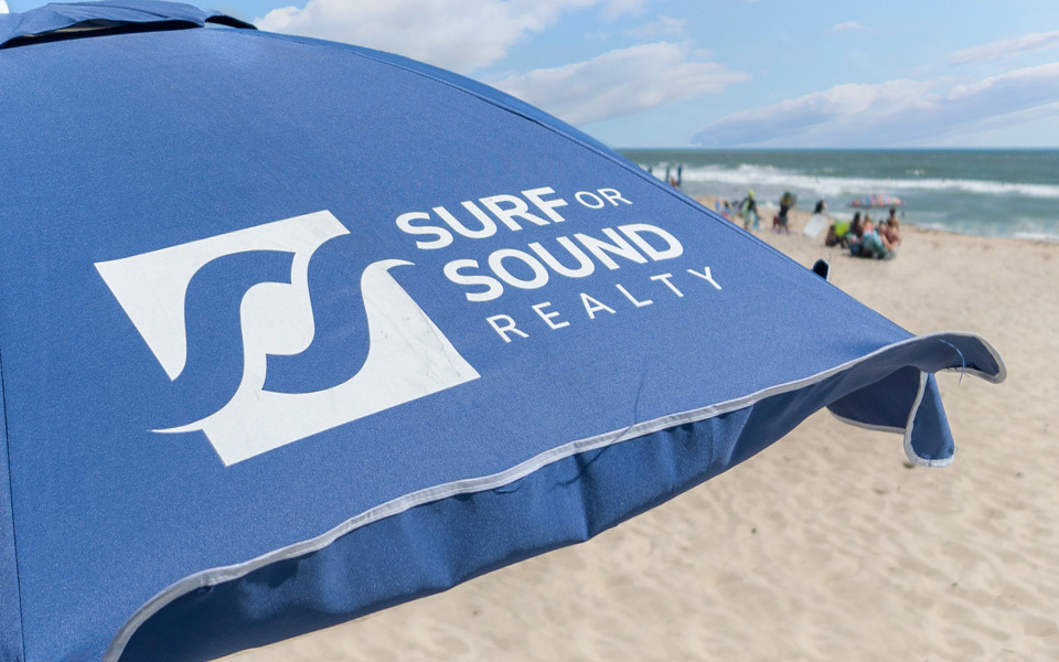 Closely cropped blue beach umbrella on a beach with the Surf or Sound Realty logo printed in white 