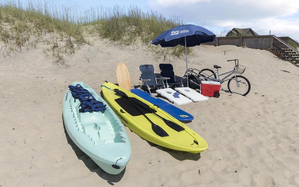 Two kayaks, two beach chairs, a beach umbrella, a cornhole game, a cooler and a bike arranges on a sandy beach