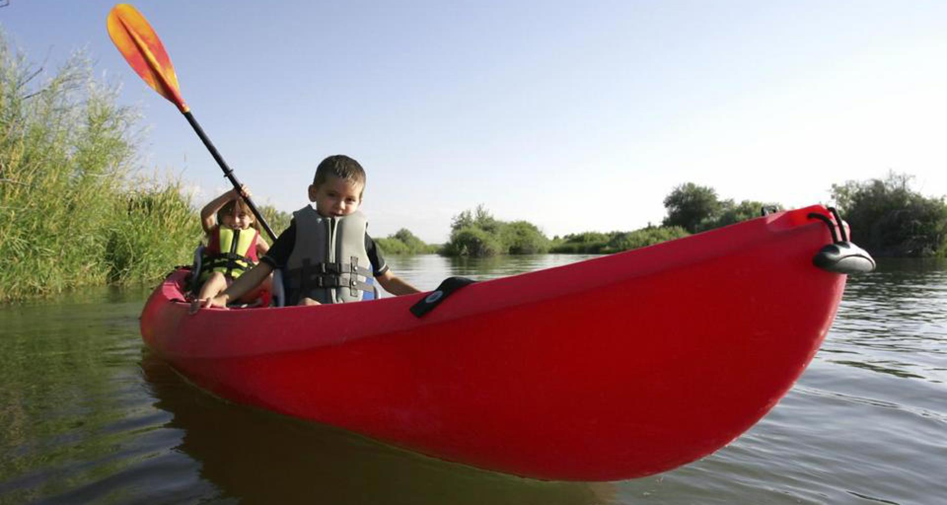 Wide angle from the bow of a bright red kayak paddled by two small kids in life jackets on the sound near marsh grasses