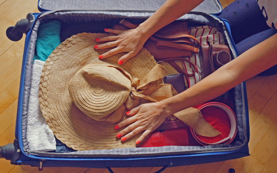 Inside of a blue suitcase seen from above with a woman's hands with red fingernails packing a beach hat