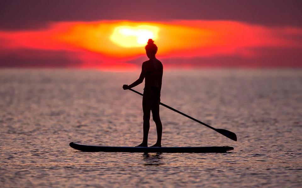 Lone paddleboarder stands on her SUP, paddle in hand, silhouetted against a rich pin and orange setting sun over the sound