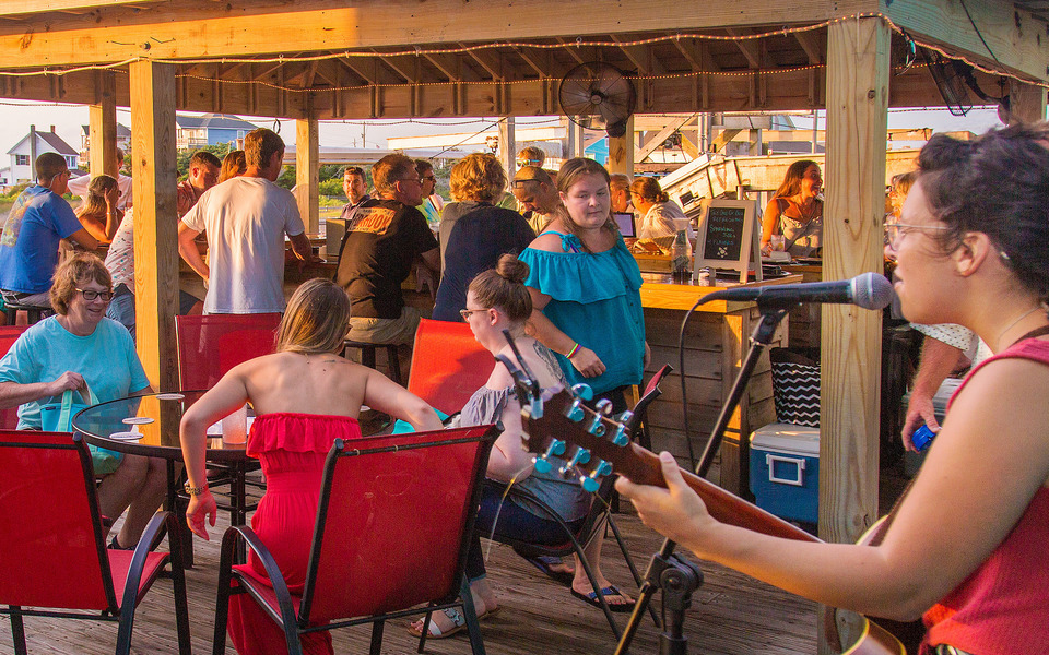 Lively outdoor bar scene with folks at table and the bar.  A singer plays guitar and sings in the foreground.
