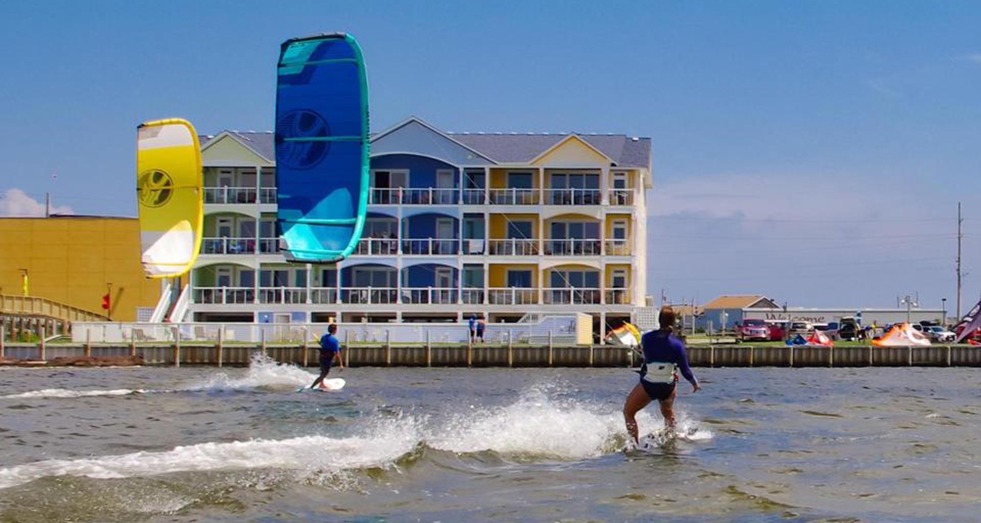 2 kiteboarders from behind as their yellow & blue kites cross in front of an colorful soundfront condo building in Rodanthe