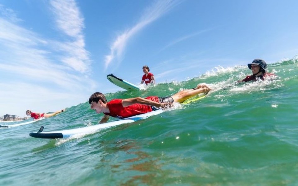 Water view of a surf instructor pushing a boy on a surfboard into a green wave under a blue sky as part of a surf lesson