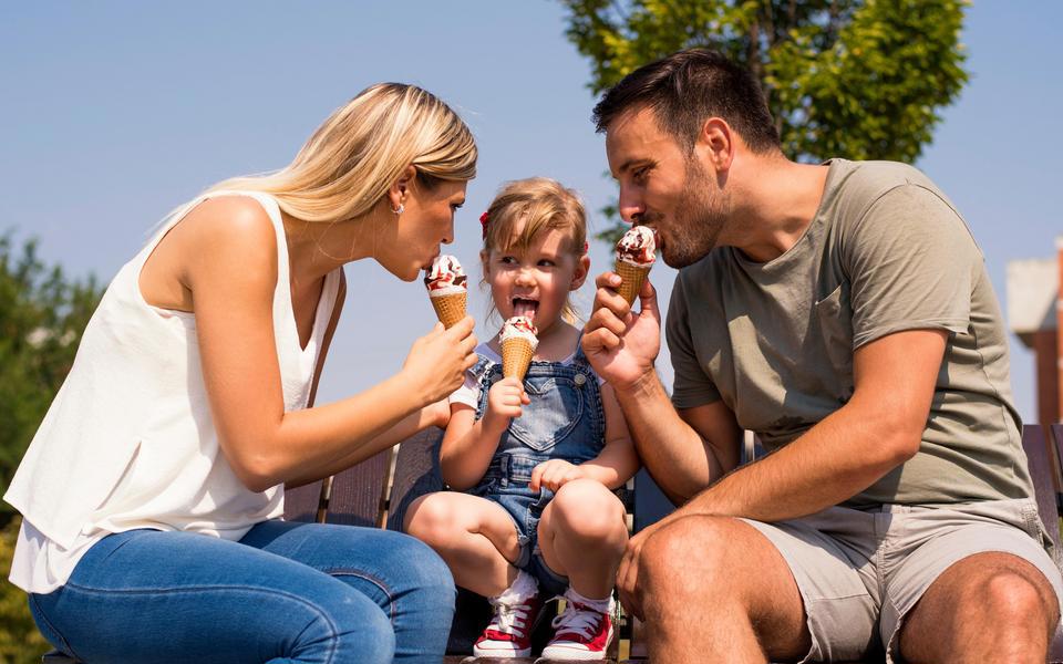 A couple flanks their young child on a bench outside.  All three lick ice cream cones under a deep blue sky.