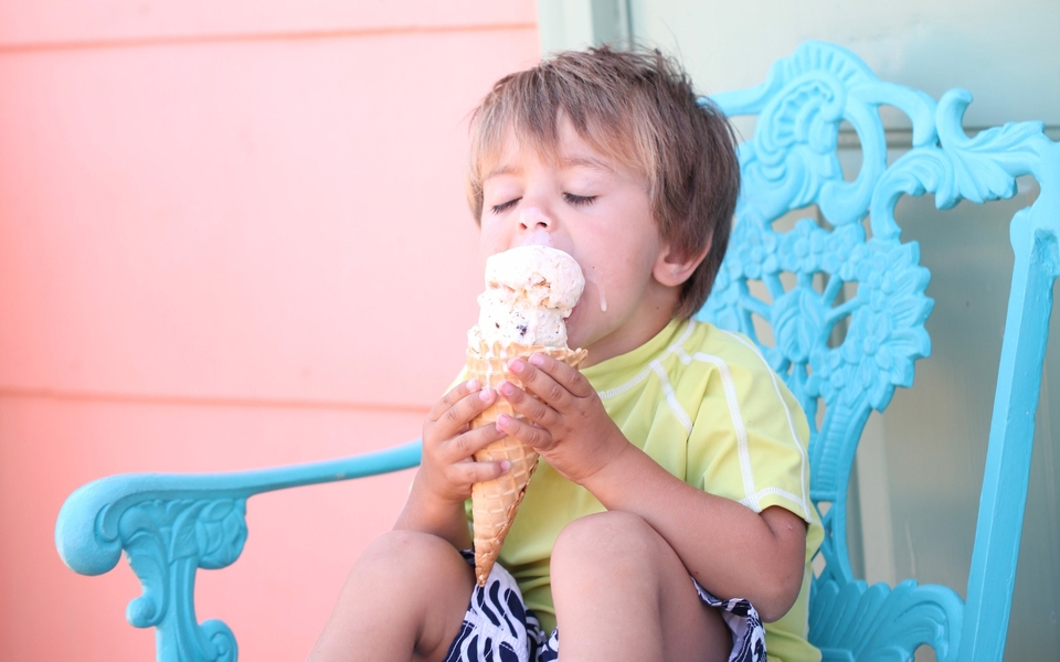 A young child eagerly licks a large ice cream cone while sitting in a light blue chair by a pink wall.