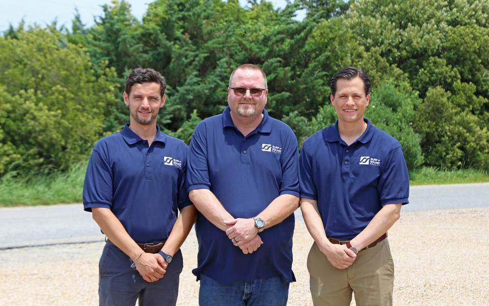 Three employees in matching blue Surf or Sound Realty polo shirts pose with hands clasped in front of green trees