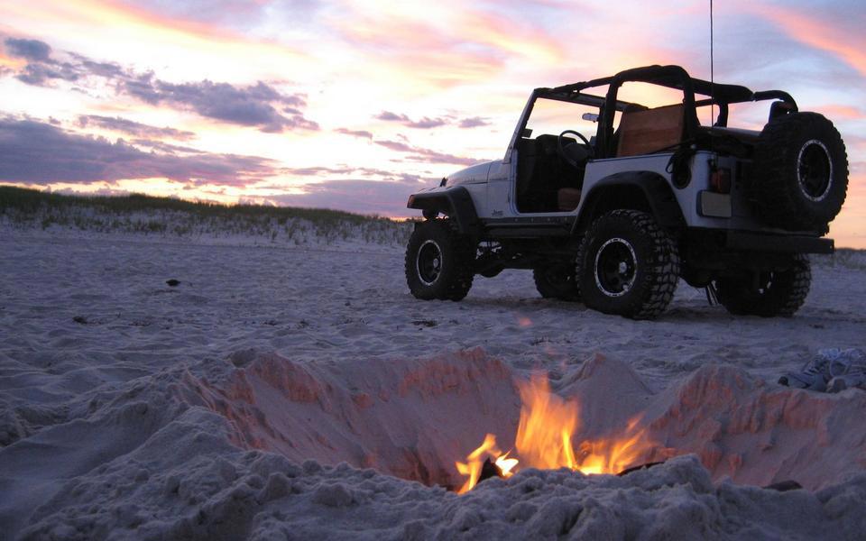 A small beach bonfire burns on sandy beach bathed in purple twilight in front of a doorless, roofless Jeep