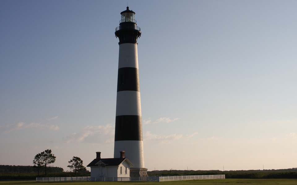 Bodie Island Lighthouse against a blue sky lit by early morning sun from behind and to the right