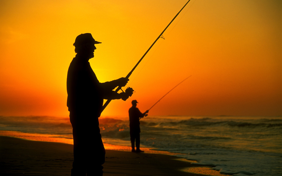Two men in hats are silhouetted by the rising sun as they fish in the ocean from as south facing beach off Hatteras Island