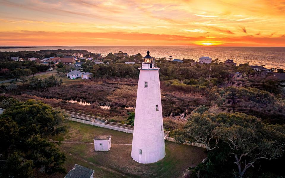 Full length of the Ocracoke Lighthouse from a drone framed by vacation homes and an orange sun setting over the Pamlico Sound