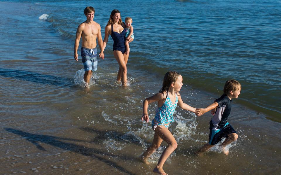 A family walks ankle-deep in gently lapping waves. Parents and two of the three kids hold hands.