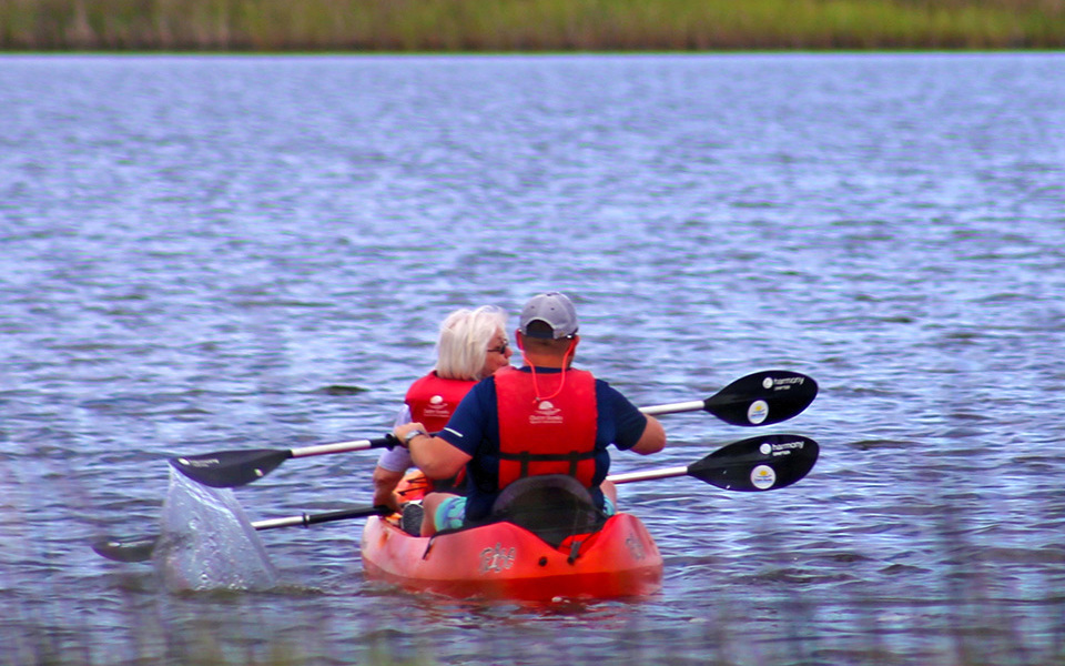 Older couple paddles away from the camera in a red tandem kayak in red life vests across the sound toward marsh grasses