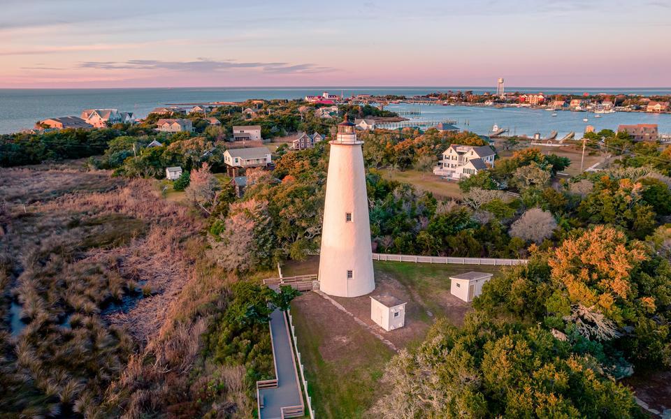 Pink light of a sunset glows over a drone view of the Ocracoke Lighthouse with the harbor and marina in the background