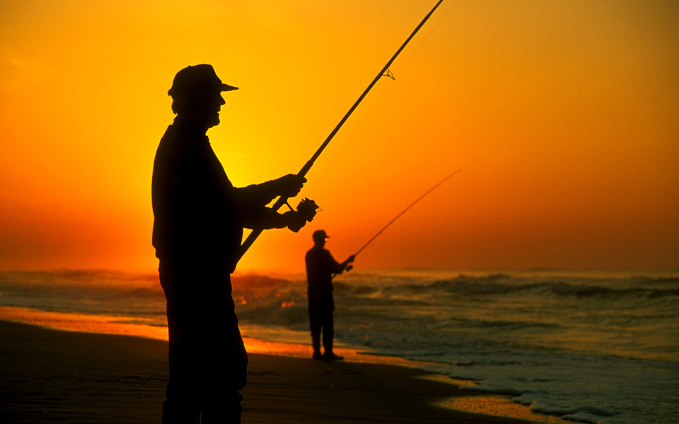 Silhouette of two men surf fishing in the ocean framed by a deep yellow and orange sunrise sky
