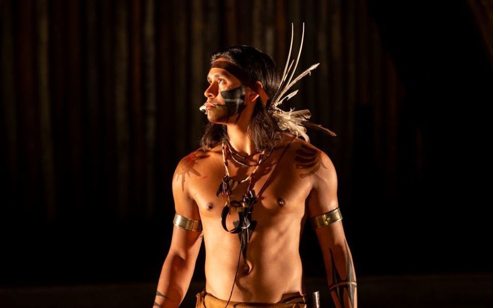 Dramatically side lit over a dark background, a promo shot of a shirtless native American character in the Lost Colony