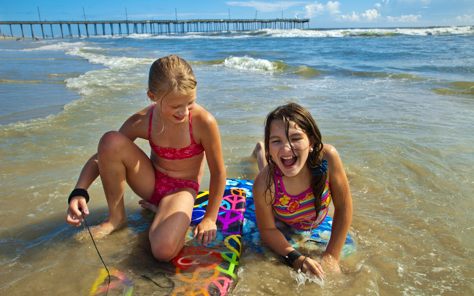 Two smiling tweens sit on colorful body boards in shallow water on a beach with a fishing pier in the background