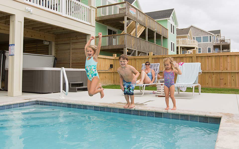 Three small children mid-jump into a private pool by a Hatteras Island vacation home as a smiling woman looks on