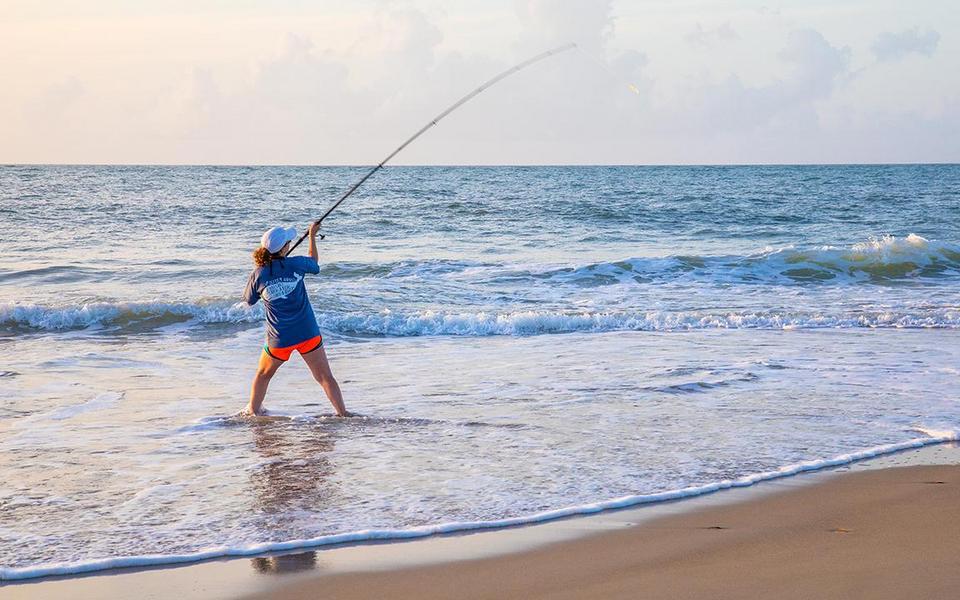 A woman surf fishes with a long fishing rod standing in shallow water of the ocean as small waves lap onto the beach