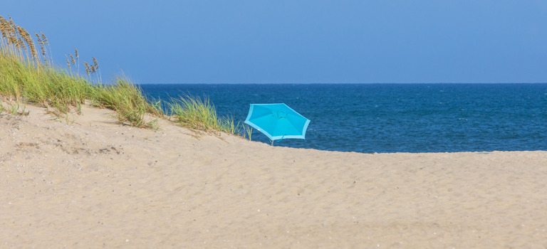 A lone light blue umbrella on a Salvo beach with a sea oat covered dune to the left and the ocean horizon to the right