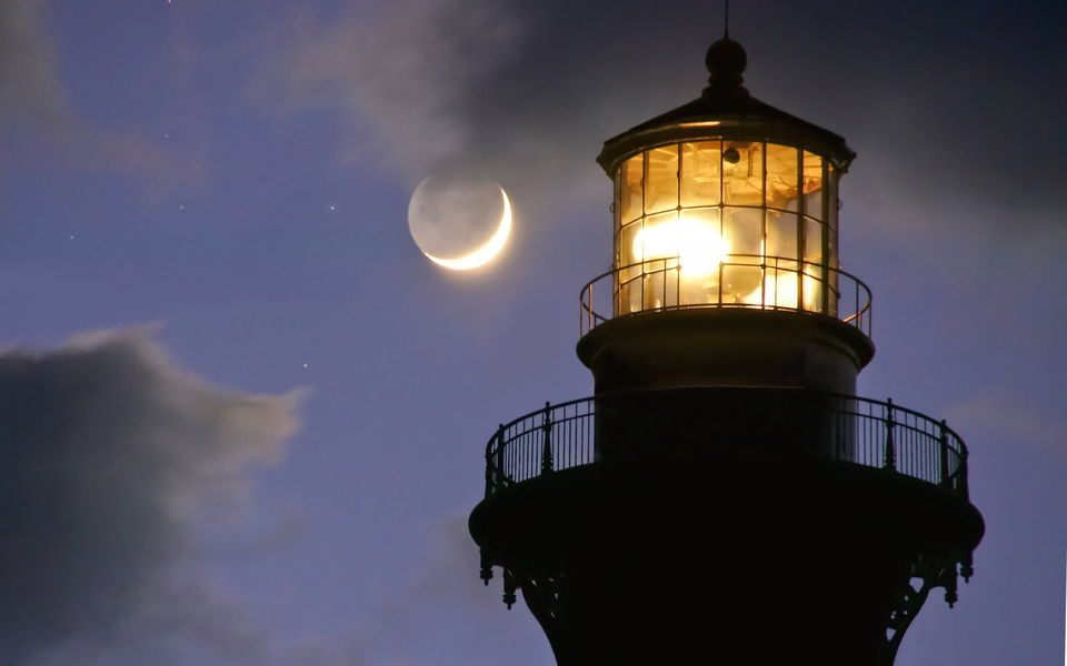 Close up silhouette of the top of the Cape Hatteras Lighthouse at night with a sliver of the moon in a purple sky