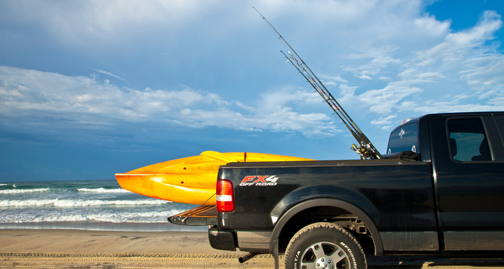 The back half of a black pickup truck with a yellow kayak and some fishing poles parked on the beach by the ocean