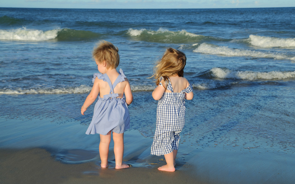 Two toddlers in blue and white jumpers stand at the edge of the shore facing the ocean on a beach on Hatteras Island, NC