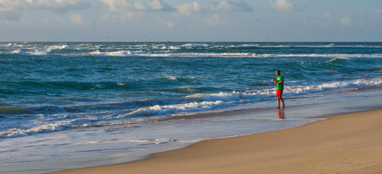 A lone man in red shorts and a light green shirt surf fishes, casting into the ocean on a stretch of beach in Frisco, NC