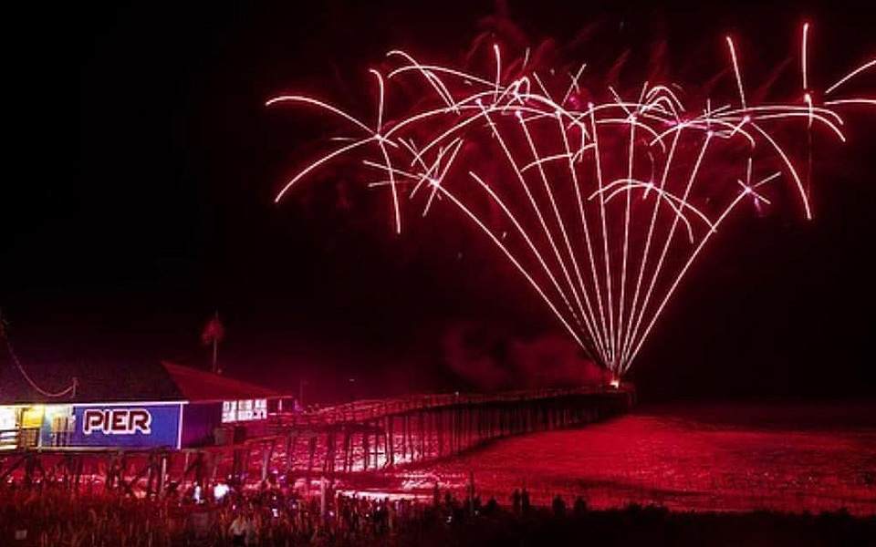 Pink fireworks shoot up into a night sky from the end of the Avon Pier over the ocean as crowds look on